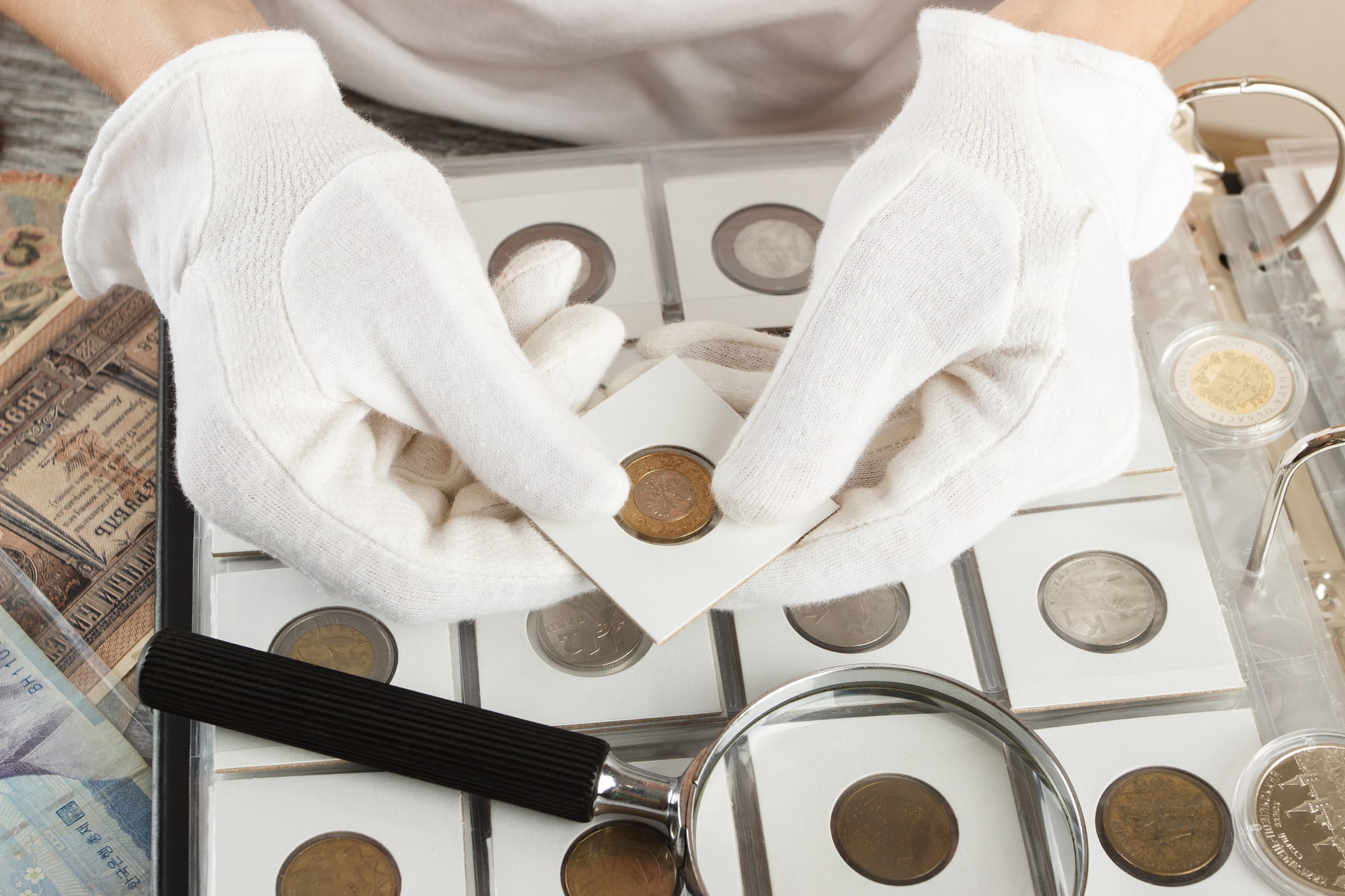 A person in white gloves examines a box of coins with a magnifying glass, focusing on the details of the coins.