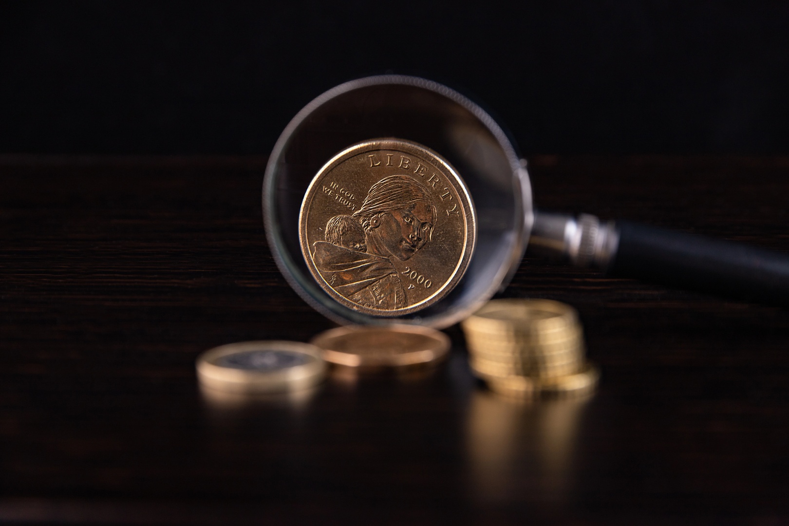 A gold coin and several coins are arranged on a table beside a magnifying glass, highlighting their details.